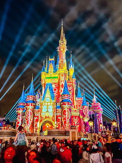 Cinderella Castle lit up with vibrant colors and spotlights beaming into night sky with crowd watching