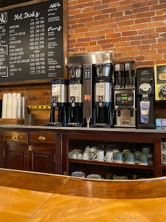 Coffee shop counter with coffee machines, cups, menu boards, and a brick wall