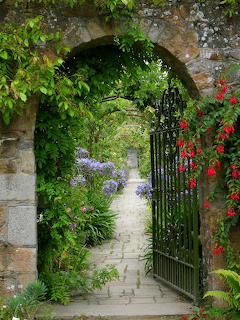 Stone arch with black iron gate leading to garden path surrounded by plants and flowers
