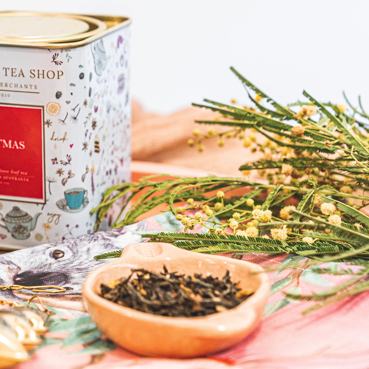 Tea tin beside a wooden spoon filled with loose tea leaves and yellow flowering branches