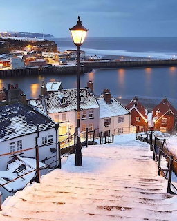 Snowy steps with a streetlamp overlooking village rooftops, harbor, and sea at dusk