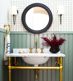 Powder room with marble sink, gold fixtures, sage green wainscoting, round black mirror, and red plant