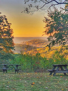 Picnic tables at scenic overlook with autumn trees framing golden sunset over hills