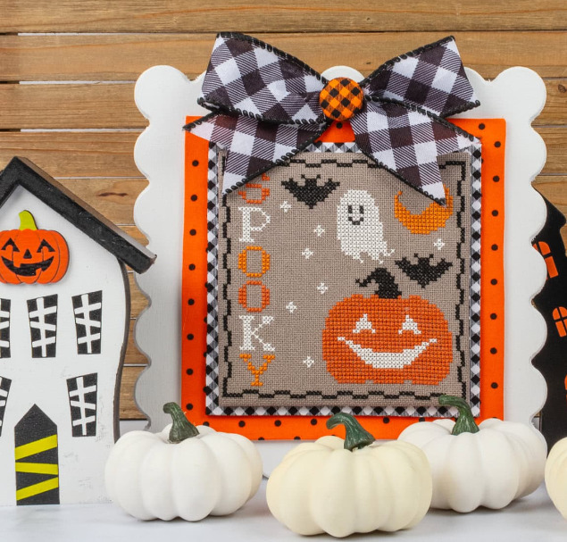 Framed Halloween cross stitch with ghost, pumpkin, and BOO lettering, decorated with checkered bow and white pumpkins