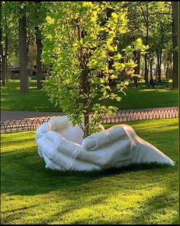 White sculpture of large hands cradling a young tree on a park lawn with green trees in the background