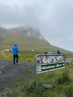 Person in blue jacket walking past Múlafossur 300m directional sign with fogy green mountains behind