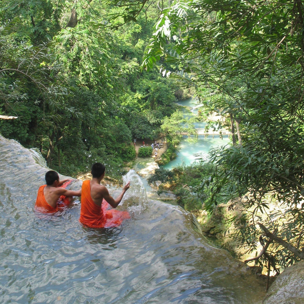 Two people in orange vests sitting in a shallow waterfall pool surrounded by lush forest