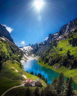 Turquoise alpine lake between steep green mountains with a chalet below and bright sun overhead