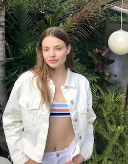 Young woman with long brown hair wearing white denim jacket and striped crop top among green plants