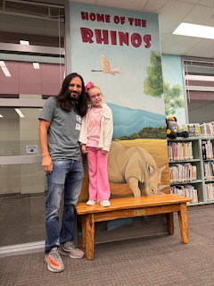 Man and girl standing beside cardboard rhino on wooden bench in school library