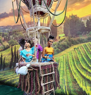 Two dolls in colorful outfits standing in hot air balloon basket over vineyard at sunset