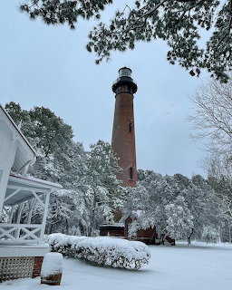 Red brick lighthouse in snow with bare trees and white building