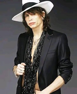 Man in white hat and black blazer with beaded necklaces posing against gray backdrop