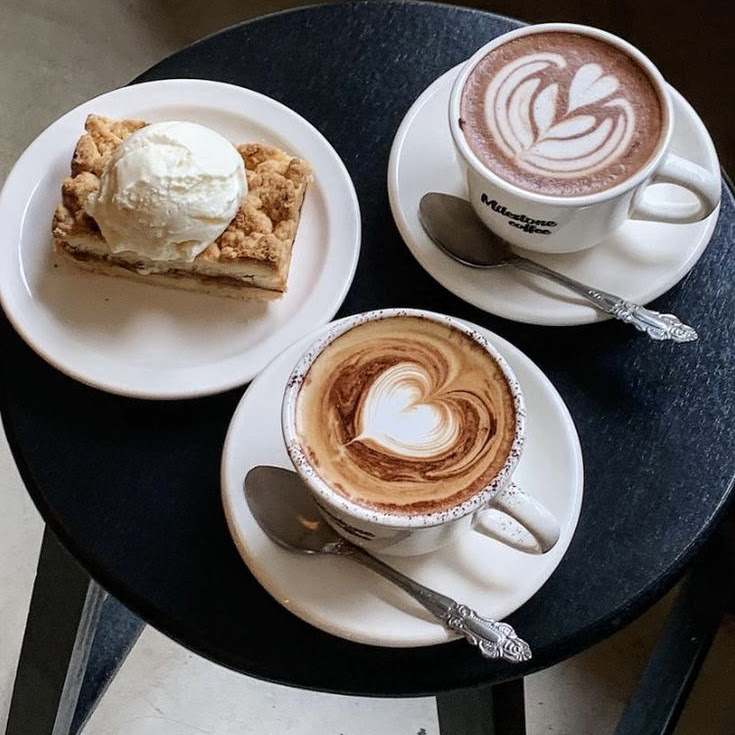 Two cappuccinos with latte art and a slice of apple crumble topped with ice cream on a table