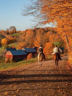 Two people riding horses down a dirt road lined with colorful fall trees and red barns in Vermont