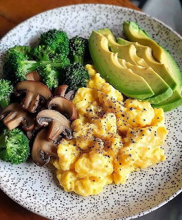 Breakfast bowl with scrambled eggs, avocado slices, broccoli florets, and mushrooms on white speckled plate