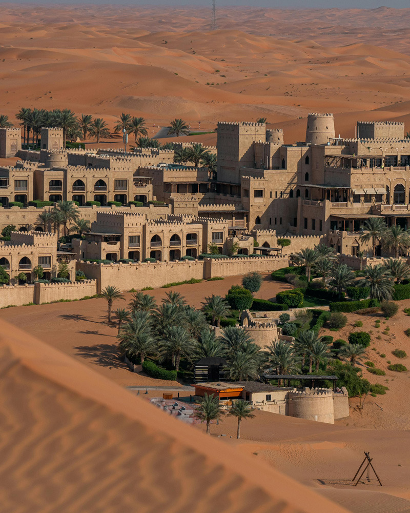 Aerial view of a desert resort with beige buildings and palm trees surrounded by rolling sand dunes