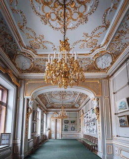 Ornate palace hall with gilded ceiling, chandelier, tall windows, and framed art