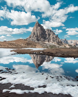 Jagged Dolomites mountain peak reflected perfectly in calm alpine lake water with snow patches and blue sky