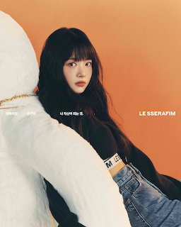 Young woman with long dark hair posing beside a giant white teddy bear on an orange background.
