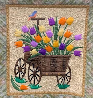 Quilt artwork showing wooden cart with orange and purple tulips and bluebird