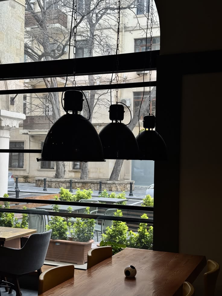 Three black pendant lights hanging in front of an arched cafe window with city buildings and trees outside