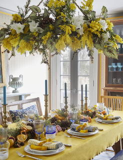 Dining table with yellow tablecloth, candles, citrus and a floral chandelier with mimosa and white flowers