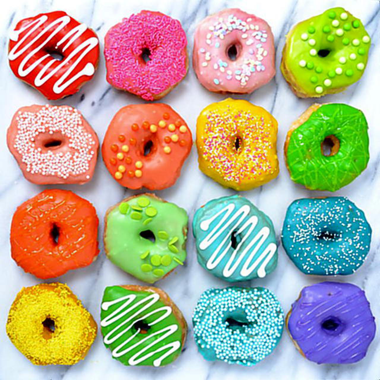 Sixteen colorful glazed donuts arranged in a 4x4 grid with various toppings and bright colors on marble surface