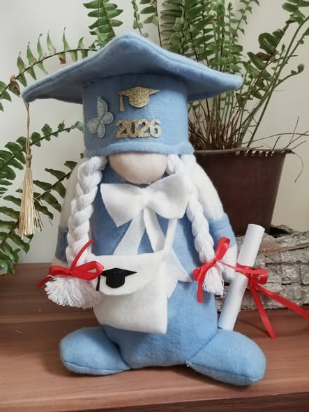 Blue fabric graduation gnome with cap, gown, braids, and diploma on a wooden shelf with plants