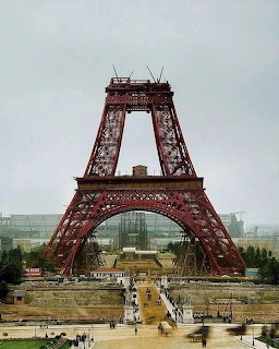 Colorized photo of the Eiffel Tower under construction with scaffolding and red-painted iron framework