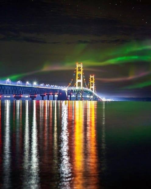 Illuminated suspension bridge at night with green and purple northern lights reflecting on water
