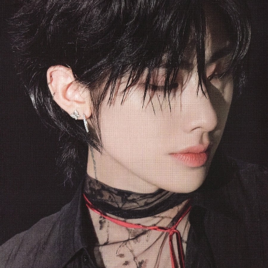 Portrait of person with black hair and pale makeup wearing dark clothing and neck accessories