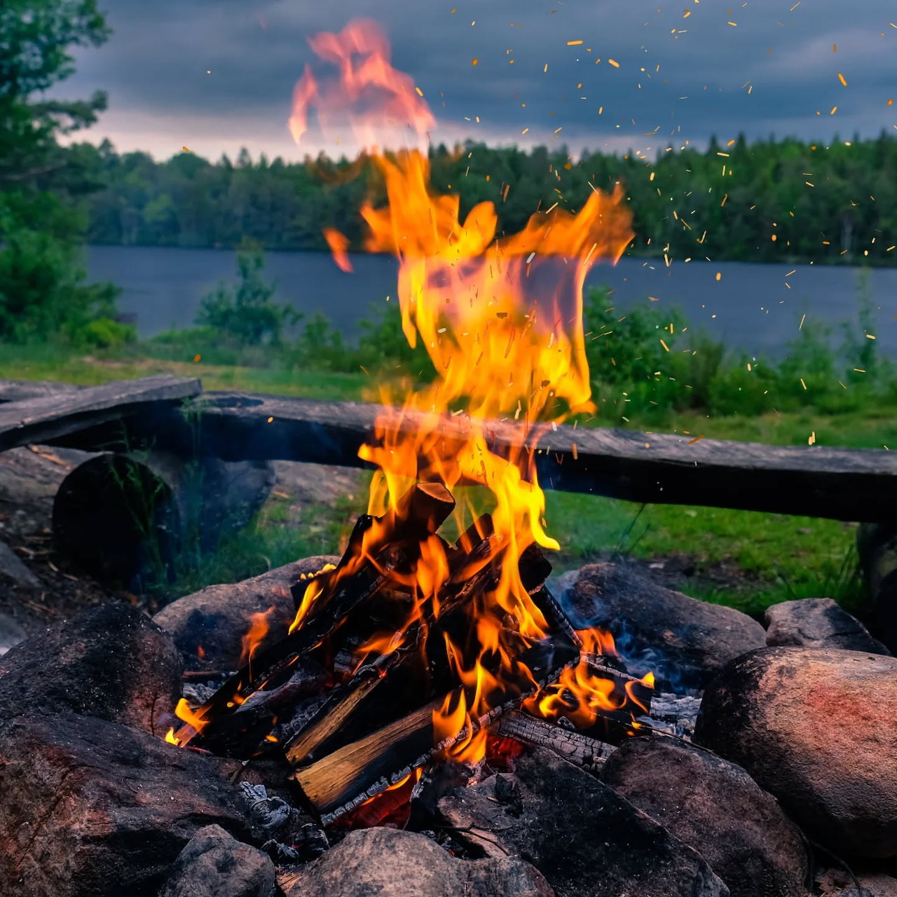 Campfire burning in a stone fire pit beside a lake at dusk