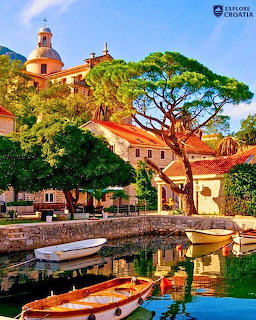 Mediterranean village with red-roofed buildings, church tower, harbor boats, and large pine tree