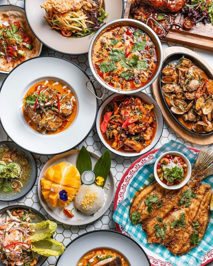 Overhead view of multiple Thai dishes including curries, clams, grilled fish, and mango sticky rice on a table