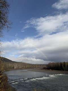 River flowing through a forested valley with mountains and a faint rainbow in a cloudy blue sky