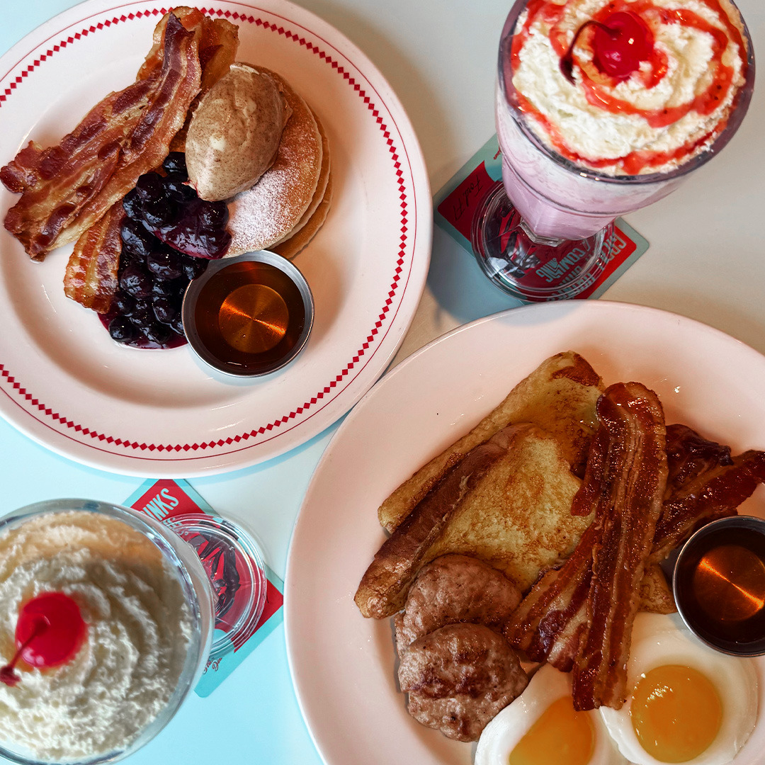 Overhead view of breakfast plates with bacon, eggs, toast, pancakes, whipped cream, and shakes