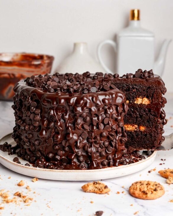 Round chocolate cake densely covered with chocolate chips on white plate, cookies and teapot nearby