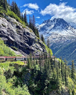 Railway track curves along rocky cliff with snow-capped mountains and evergreen trees in Alaska