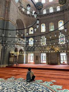 Person praying inside a grand mosque with domed ceiling, stained glass windows, and red carpet