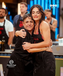 Two women hugging on MasterChef Celebrity TV set with kitchen background and other contestants