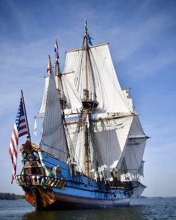 Blue tall ship with white sails and American flag sailing on calm water under blue sky