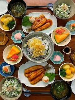 Top-down Japanese meal with grilled fish, noodles, rice, soup, fruit, and side dishes