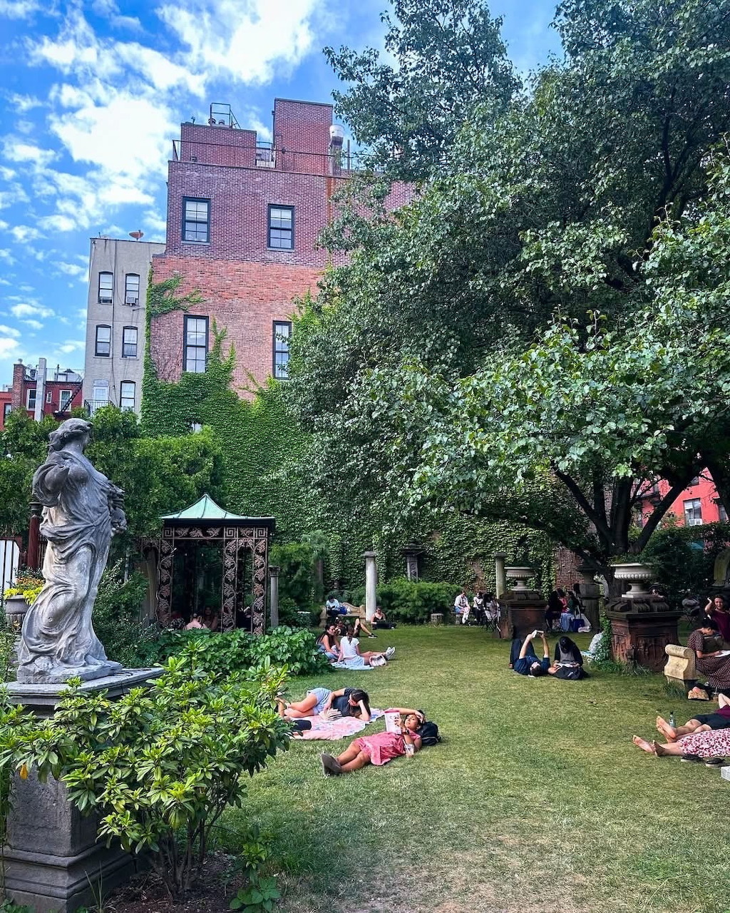 People relaxing on grass in small urban park surrounded by trees and brick buildings in Nolita, NYC
