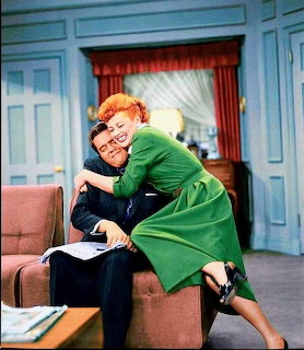 Lucille Ball in green dress hugging Desi Arnaz on I Love Lucy set with blue walls and red curtain backdrop