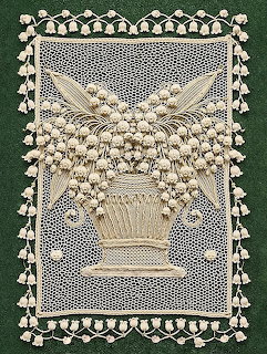 Detailed white lace piece showing lilies of the valley in a woven basket with ornate lace border on green fabric