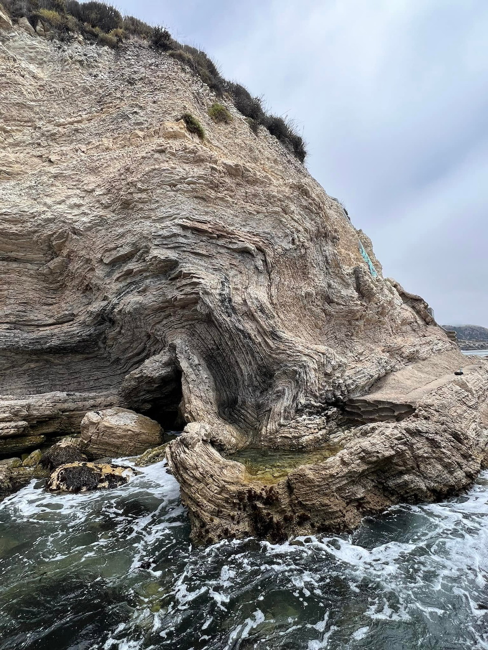 Tall weathered coastal cliff with stratified rock layers and cave opening above churning ocean water