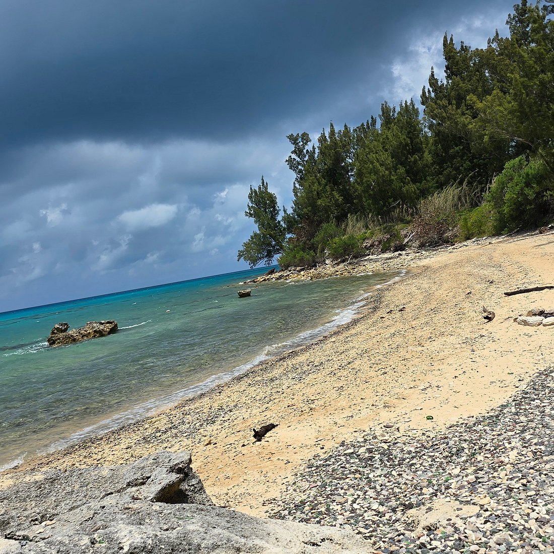 Sandy beach with turquoise water, rocks, green hillside vegetation, and stormy clouds