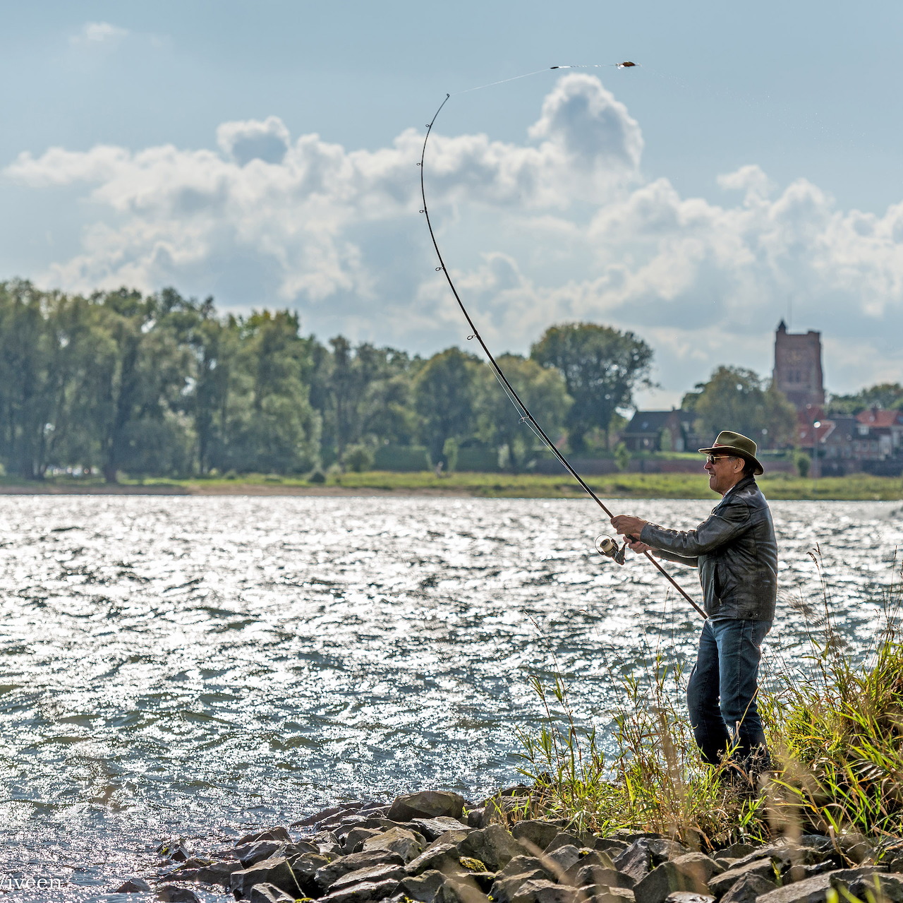 Fisherman casting fishing rod on River Waal with Woudrichem town visible across the water under cloudy sky