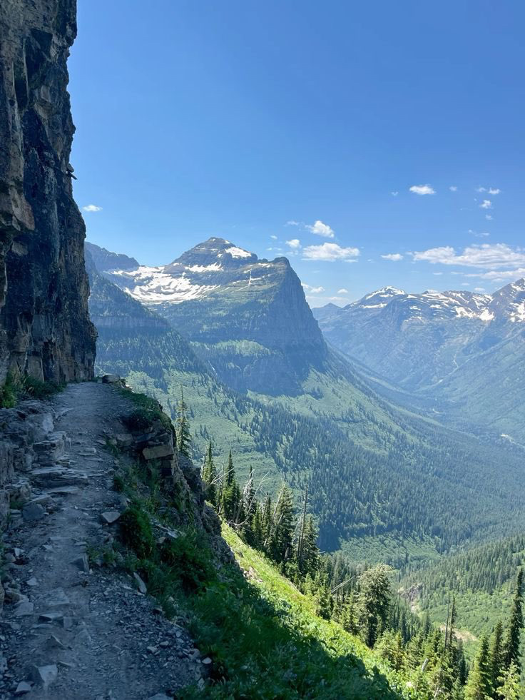 Narrow hiking trail along a rocky cliff with green valley and snow-capped mountains in the background